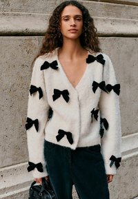 Weiße, flauschige Strickjacke mit mehreren schwarzen Samtbows, die auf den Stoff genäht sind, tiefem V-Ausschnitt und langen Ärmeln. Getragen mit dunklen Hosen.
