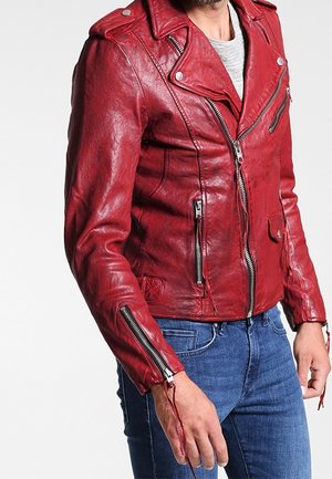 Uomo che indossa una giacca biker in pelle rossa con tasche e maniche con cerniera sopra una camicia bianca e jeans blu, in piedi contro uno sfondo bianco.