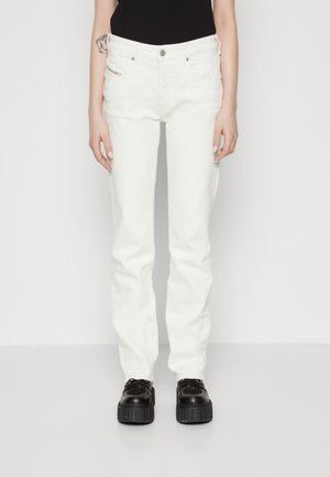 Persoon die witte rechte jeans, zwarte platformschoenen en een zwart topje draagt, staand tegen een effen witte achtergrond.