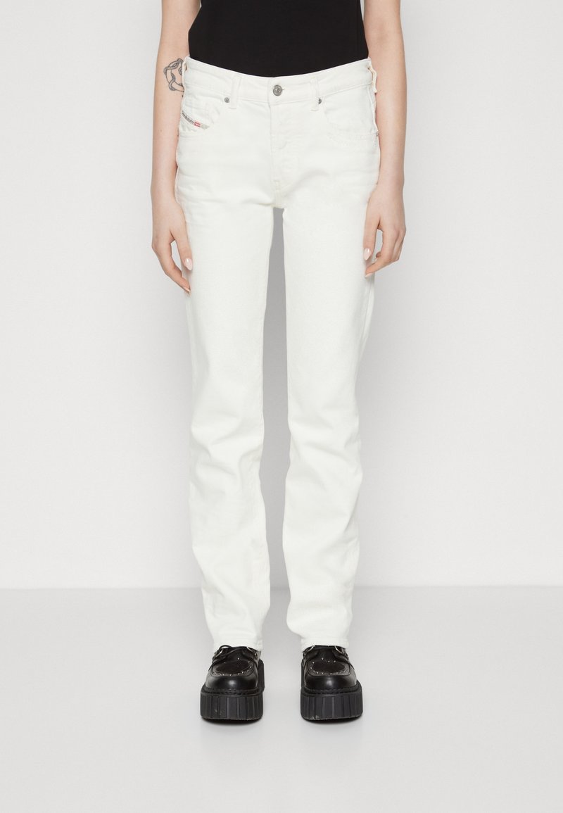 Persoon die witte rechte jeans, zwarte platformschoenen en een zwart topje draagt, staand tegen een effen witte achtergrond.