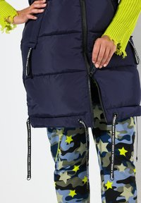 Marinblå pufferväst med dragkedja, med hög krage och dragkedjefickor. Matchad med leggings i kamouflagemönster i grönt och blått.