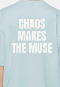 Camiseta de color azul claro con texto blanco en negrita en la parte de atrás que dice "EL CAOS HACE A LA MUSA". Cuello redondo y mangas cortas.