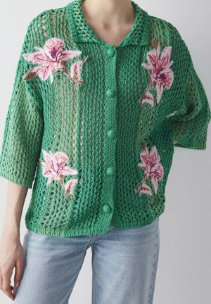 Green open-knit cardigan with pink floral embroidery, buttoned front, loose sleeves, worn with light blue jeans.