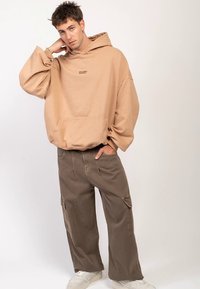Beżowa, oversize'owa bluza z kapturem z przednią kieszenią i ściągaczami przy mankietach, w połączeniu z luźnymi brązowymi spodniami cargo i białymi trampkami.