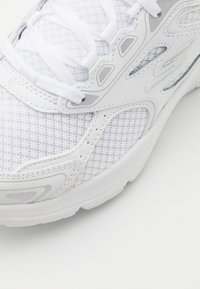 Chaussure de sport blanche avec une tige en mesh respirant, des accents synthétiques, une semelle texturée et des lacets plats. Comprend des détails gris subtils.