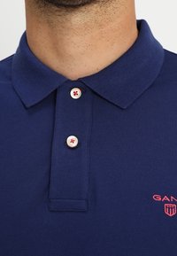 Marinblå polo skjorta, i bomull, klassisk krage, tre-knapplist med vita knappar som har röda detaljer, broderad logotyp på bröstet.