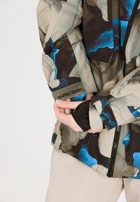 Wasserabweisende Jacke in Beige mit blauen und schwarzen abstrakten Mustern, ausgestattet mit einem Reißverschluss, W-PRO 10000-Label und strukturiertem Stoff.