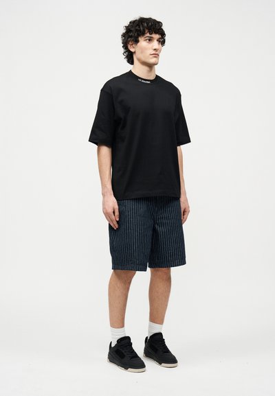 Άνδρας με σγουρά μαλλιά στέκεται φορόντας μια μαύρη φαρδιά μπλούζα, κοντά σορτς με ρίγες μέχρι το γόνατο, λευκές κάλτσες και μαύρα παπούτσια πάνω σε λευκό φόντο.