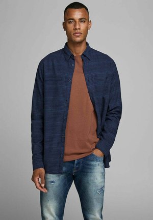 Camicia a righe navy con bottoni sopra una maglietta marrone, abbinata a jeans blu strappati. La camicia presenta un design casual e tasche a toppa.