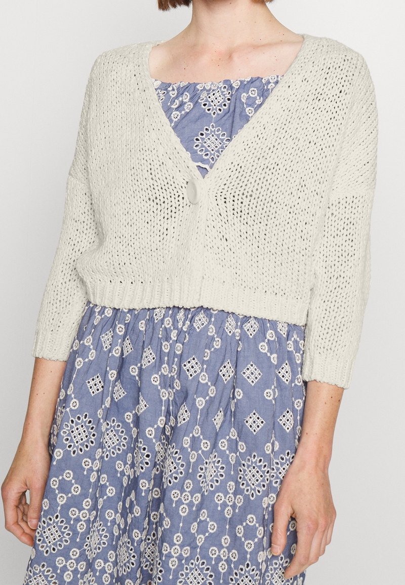 Off-White Strickjacke mit V-Ausschnitt, dreiviertellangen Ärmeln und einem einzigen Knopfverschluss, getragen über einem gemusterten blauen Kleid.