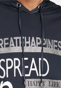 Marinblå hoodie med silverglittertext som säger "SPRIDA LYCKA." Har en dragsko med metalländar och en mjuk textur.