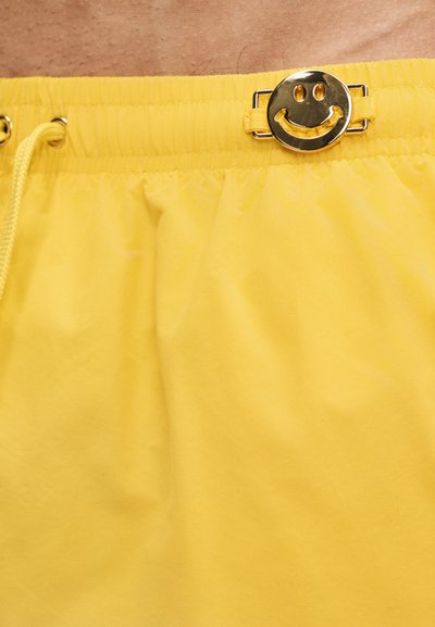 Κίτρινα σορτς με ελαστική ζώνη μέσης και μεταλλική χρυσή αγκράφα με σχέδιο χαμογελαστού προσώπου, καθώς και κίτρινο κορδόνι στη μέση ενός ατόμου.