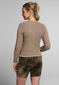 Hellbrauner Strickpullover mit einem strukturierten Zickzackmuster auf dem Rücken, gerippten Bündchen und weißem Saum. Kombiniert mit dunkelbraunen Shorts.