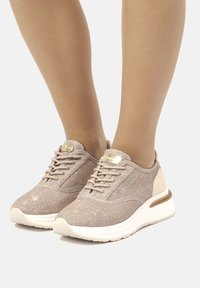 Baskets de sport beige clair avec un dessus en textile pailleté, bout rond et semelles blanches. Elles possèdent des lacets et un subtil accent métallique sur le talon.