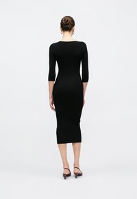 Γυναίκα με μαλλιά σε σινιόν, φοράει εφαρμοστό μαύρο ριμπ μέσο φόρεμα με μανίκια τριών τετάρτων και μπορντό πέδιλα με χαμηλό τακούνι και λουρί πίσω.