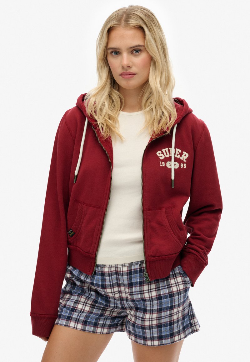 Sudadera roja con cremallera y cordones, que presenta un logo y bolsillos frontales. Combinada con pantalones cortos de cuadros en azul, blanco y rojo.
