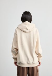 Pessoa vista de costas, usando um sweatshirt com capuz bege e calças cargo castanhas, contra um fundo claro e simples.