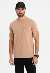 Ljust beige kortärmad t-shirt med ficka, rund halsringning och en jämn textur. Har en liten logotyp på fickområdet.