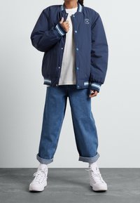 Granatowa bomberka z paskami na mankietach i kołnierzu, z niebieskimi zatrzaskami i okrągłą naszywką z logo. Noszona na białej koszuli i niebieskich dżinsach.