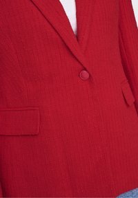 Rød tekstureret blazer med hakket revers, enkelt knaplukning og to forlommer. Stoffet har et ribbet mønster for ekstra detaljer.