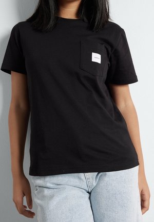 Femme portant un t-shirt noir uni à manches courtes avec une petite étiquette blanche sur la poche poitrine et un jean en denim bleu clair.