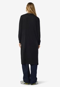 Lang svart genserkjole, mykt stoff, lange ermer, avslappet passform, bakfra, kombinert med løstsittende blå jeans og lyse sko.