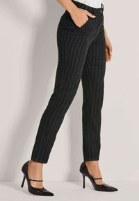 Vrouw in zwart gestreepte slim-fit broek en zwarte hoge hakken, met hand in de zak, tegen een neutrale achtergrond.