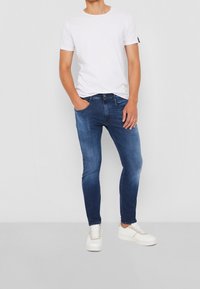 Jeans ajustados de color azul oscuro hechos de denim, combinados con una camiseta blanca de manga corta y zapatillas blancas con un acento en gris claro.