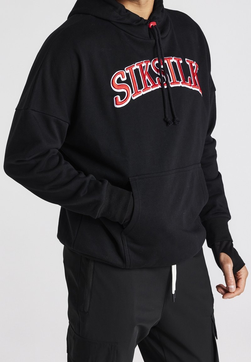 Personne portant un sweat à capuche noir SikSilk avec des inscriptions rouges et blanches, un pantalon noir, les mains partiellement visibles dans les poches et en position détendue.