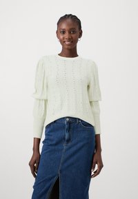 Lys grønn strikkegenser med et teksturert bølgemønster, rund hals og puffede ermer, parret med en mørk denimskjørt med slisse.