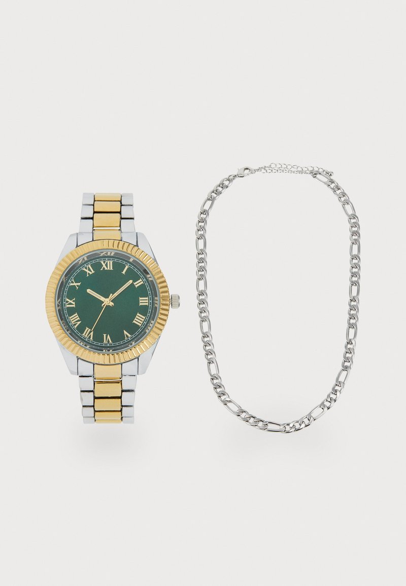 Montre aux tons doré et argenté avec un cadran vert, des chiffres romains et une lunette côtelée. Comprend un collier en chaîne argentée à côté.
