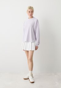 Camicia a maniche lunghe viola chiaro con scollo rotondo, abbinata a una gonna bianca plissettata. Il modello indossa sneakers bianche e calzini alla caviglia.