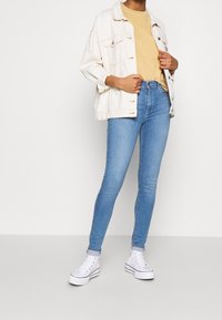 Chaqueta de mezclilla crema de gran tamaño sobre una camiseta amarilla, combinada con jeans ajustados de color azul claro y zapatillas blancas altas. Diseño simple, aspecto casual.
