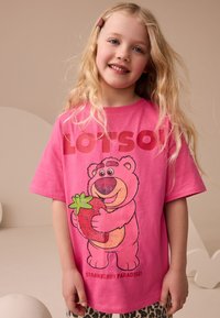 Ružové bavlnené tričko s kresleným medveďom držiacim jahodu a veľkým nápisom „LOTSO“ a pod tým „STRAWBERRY PARADISE!“.