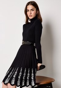 Zwarte ribgebreide jurk met lange mouwen en een hoge halslijn, voorzien van witte bloemenborduursels aan de zoom en een zwarte riem.