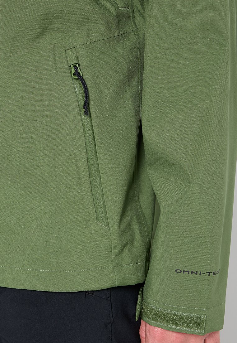 Groene waterdichte jas met een ritszak aan de zijkant, textuurstof, verstelbare manchetten en het "OMNI-TECH" logo aan de onderzijde van de mouw.