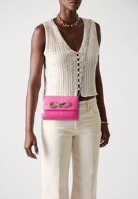 Borsa a tracolla in pelle rosa con catena, dotata di un dettaglio a nodo in argento sul davanti; indossata con un top in maglia crema e pantaloni beige chiaro.