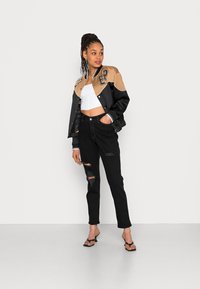 Casaco varsity preto e bege com lettering bordado, top cropped branco, jeans pretos desgastados e saltos altos pretos com tiras. Textura suave.