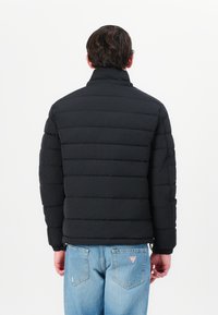 Jachetă puffer neagră cu secțiuni matlasate orizontale, guler înalt și manșete elastice, asortată cu blugi din denim albastru deschis.