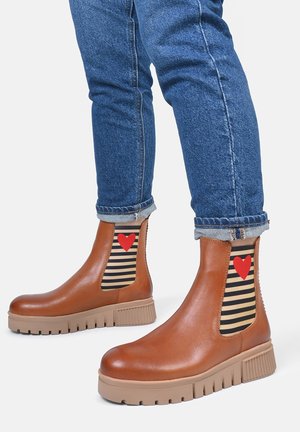 Braune Leder-Ankle-Boots mit gestreiften elastischen seitlichen Einsätzen, die ein rotes Herz zeigen, kombiniert mit hochgekrempelten Jeans und strukturierten Sohlen.