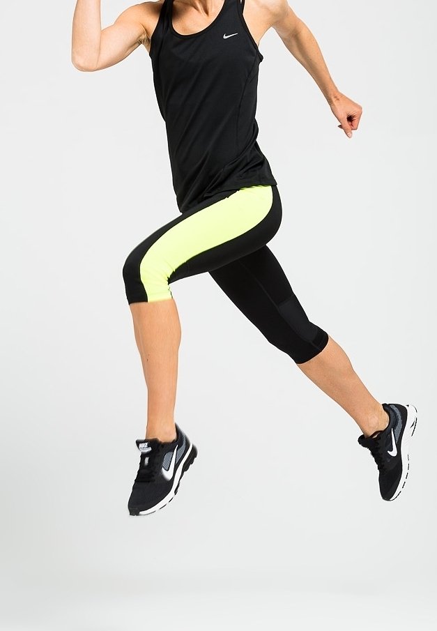 Czarna sportowa bluzka na ramiączkach z logo Nike, połączona z czarnymi legginsami 3/4 w neonowo-żółtym kolorze oraz czarnymi butami do biegania z białymi akcentami.