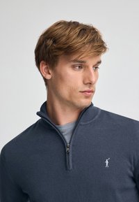 Marinblå tröja med halv dragkedja i mjukt material; har en ribbad krage och en liten broderad logotyp på bröstet.