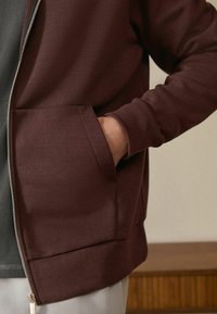 Burgunderfarbene Zip-Hoodie mit glatter Textur, seitlichen Taschen und gerippten Bündchen. Über einem grauen, gerippten Oberteil getragen, vor einem Holz-Hintergrund.