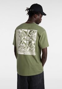 Oliivinvihreä puuvillapaita, jossa on suuri graafinen kuva takana, jossa lukee "VANS OFF THE WALL" ja ruudullisia yksityiskohtia.