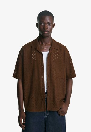 Brown short-sleeved shirt with cut-out detailing, a relaxed fit, and a pocket. Worn over a white undershirt, paired with dark jeans.