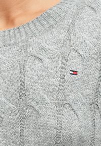 Grauer Strickpullover mit einem strukturierten Kabelmuster, ausgestattet mit einem kleinen gestickten Logo in Rot, Weiß und Blau. Rippdetail am Kragen.