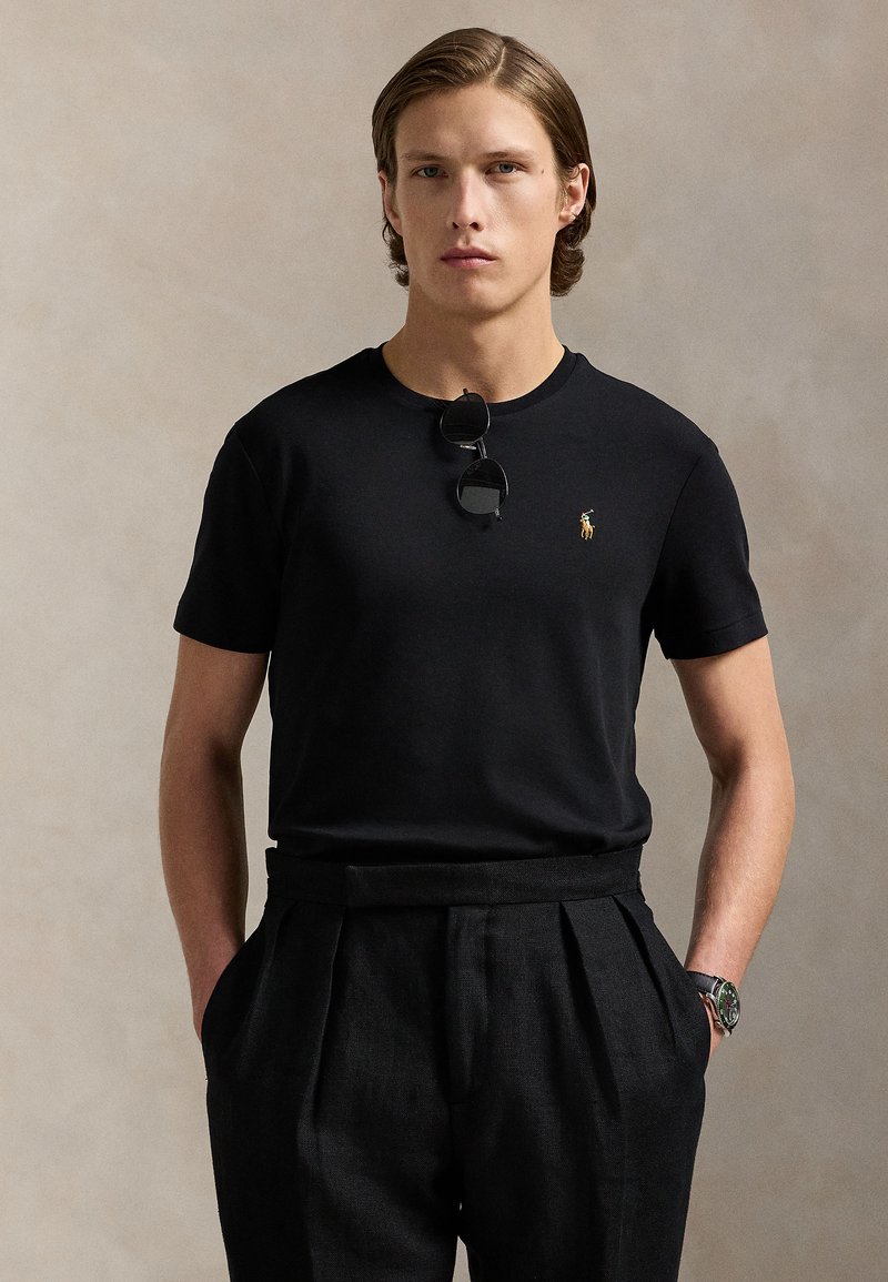 Junger Mann mit hellbraunen Haaren, der ein schwarzes T-Shirt mit kleinem Logo, schwarze plissierte Hose, Sonnenbrille am Kragen und eine Armbanduhr trägt.