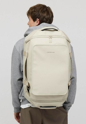 Sac à dos beige au design rectangulaire élégant, avec des bretelles réglables et une poignée supérieure. Dispose d'une poche zippée à l'avant et d'un branding discret.