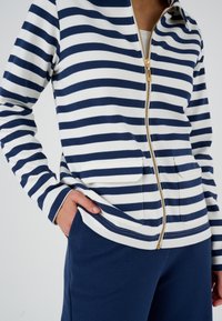 Giacca a zip a righe navy e bianche con tessuto strutturato, zip dorata e due tasche frontali, abbinata a pantaloni navy strutturati.