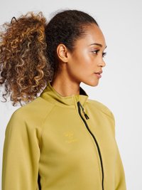 Frau mit lockigem Pferdeschwanz, die eine gelbe Jacke mit Reißverschluss und hohem Kragen trägt, schaut nach rechts vor einem schlichten hellen Hintergrund.